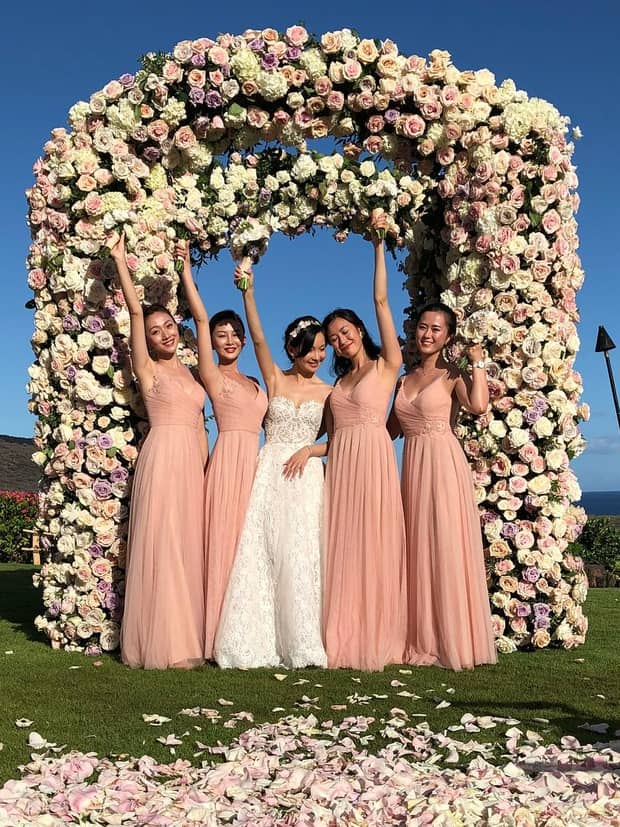 Bride and bridal party wearing pink dresses pose under wedding altar covered with roses
