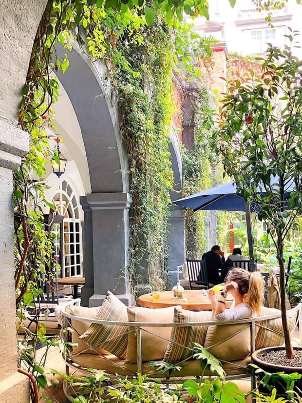 Woman drinks mimosa on curved patio sofa under grey arched doorway with vines