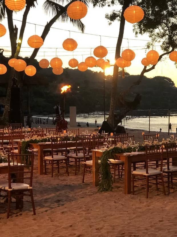 Orange paper lanterns hang over rows of dining tables on beach at sunset 