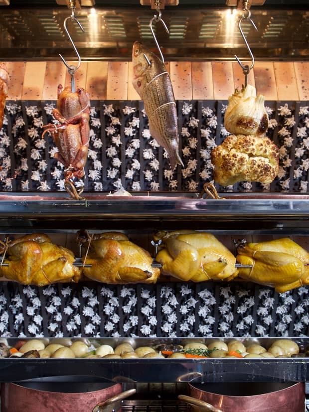 Whole chickens on rotisserie under whole fish, cauliflower hanging from hooks