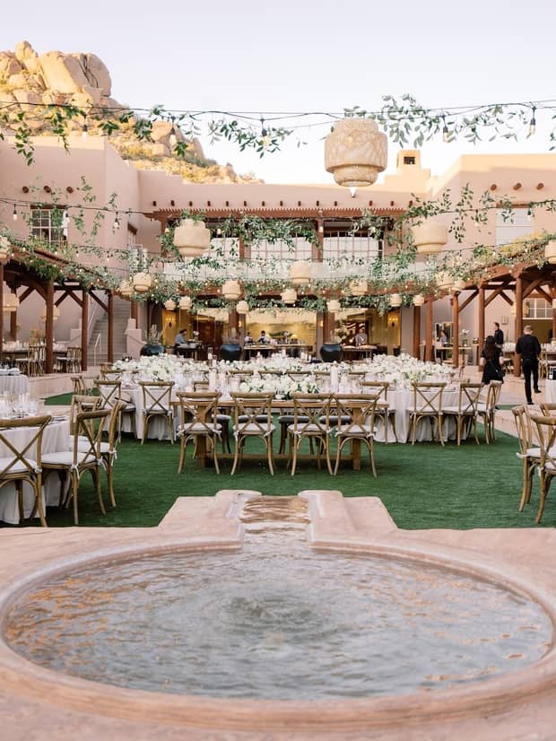 An outdoor wedding reception in a green space by a fountain, with greenery and pendant lights hanging above banquet tables with wooden chairs