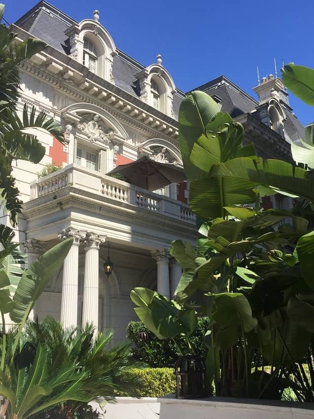 Exterior view of white pillars outside historic mansion, large tropical leaves, sunny day