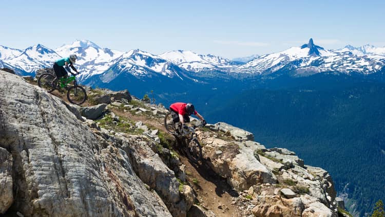 Two people on mountain bikes cycle down rocky mountain near peak, snow caps in background