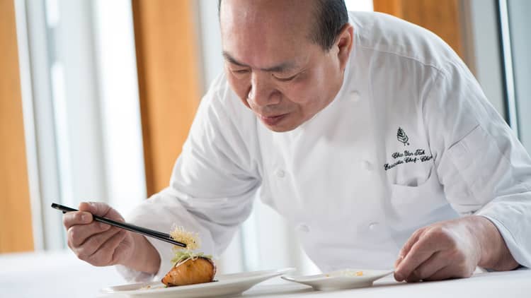 Chef in white uniform leans over grilled salmon dish on plate, adds garnish with chopsticks