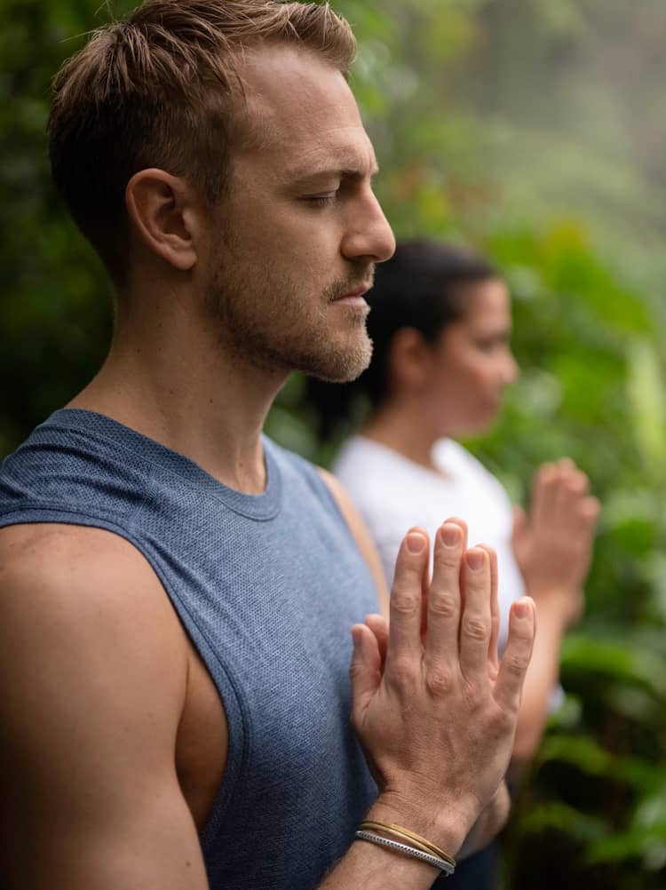 Short-haired man wearing blue sleeveless shirt stands outside with his eyes closed and his hands folded together by his chest