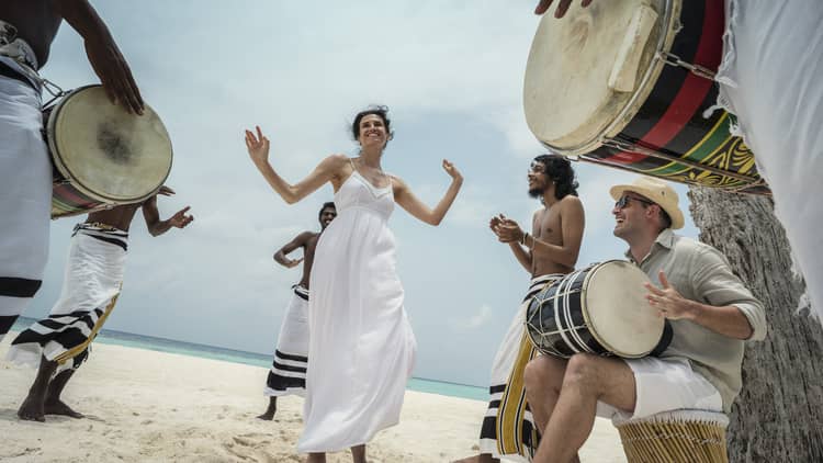 Woman wearing white dress dances in middle of drummers on beach