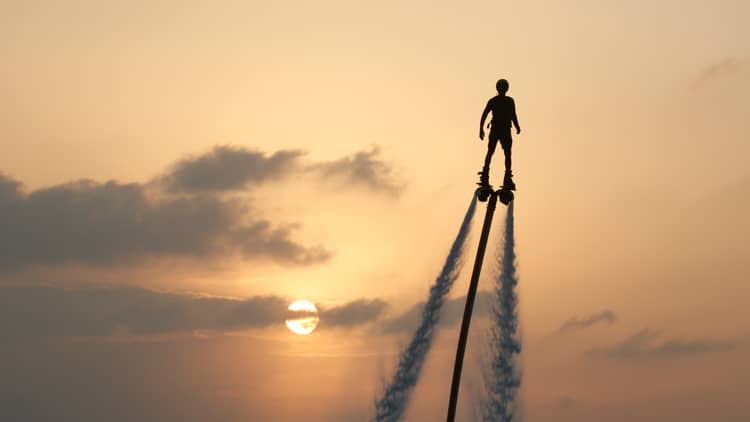 Silhouette of person soaring high above ocean on jet pack, water trails below, against sunset