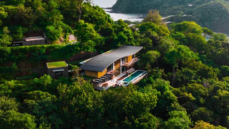 A standalone residence with a private pool sits nestled on a hillside surrounded by dense greenery with the ocean in the background