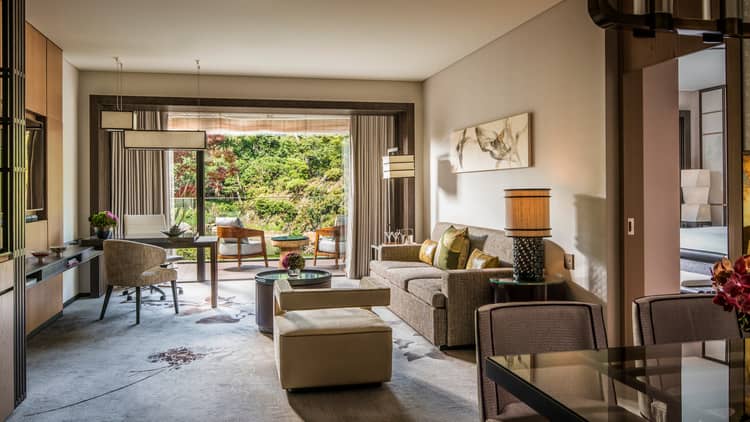 Living area with sofa, cushions, coffee table, armchair, marble desk and a private balcony with outdoor seating overlooking a lush garden