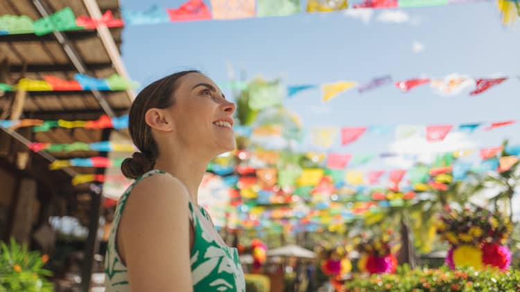 Woman with her hair pulled back in a low bun and wearing a green-and-white patterned dress stands beneath rows of colored squares hanging in an outdoor market