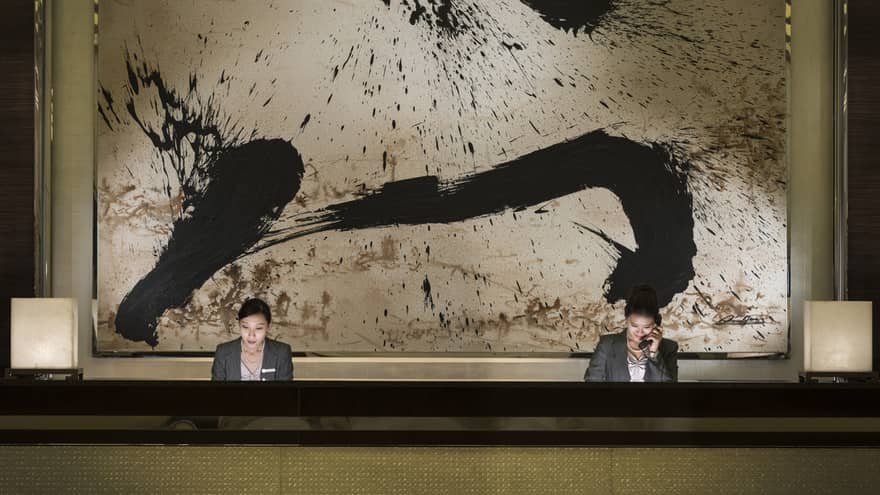 Two hotel reception staff at long desk under large black-and-white abstract painting