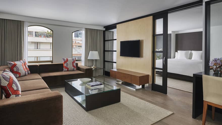 One-Bedroom Suite living room with L-shaped brown sofa, red and grey geometric accent pillows, TV wall divider to bedroom