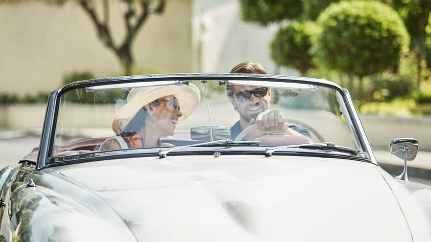 Smiling couple drive in silver luxury convertible car on sunny day