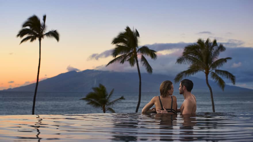 Couple embracing in an infinity pool at sunset, overlooking the ocean and distant mountains with palm trees