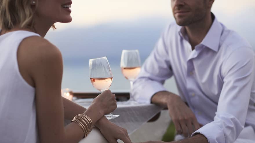 Close-up of woman and man in white, sitting and holding glasses of rose wine at dusk