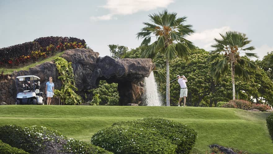 Man swings golf club on course by rock and waterfall, woman stands by cart