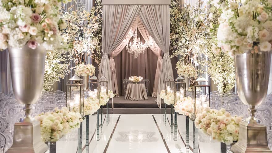 An elegantly setup wedding venue with indoor seating, featuring white and pink flowers, crystal chandeliers and champagne-coloured curtains at the altar