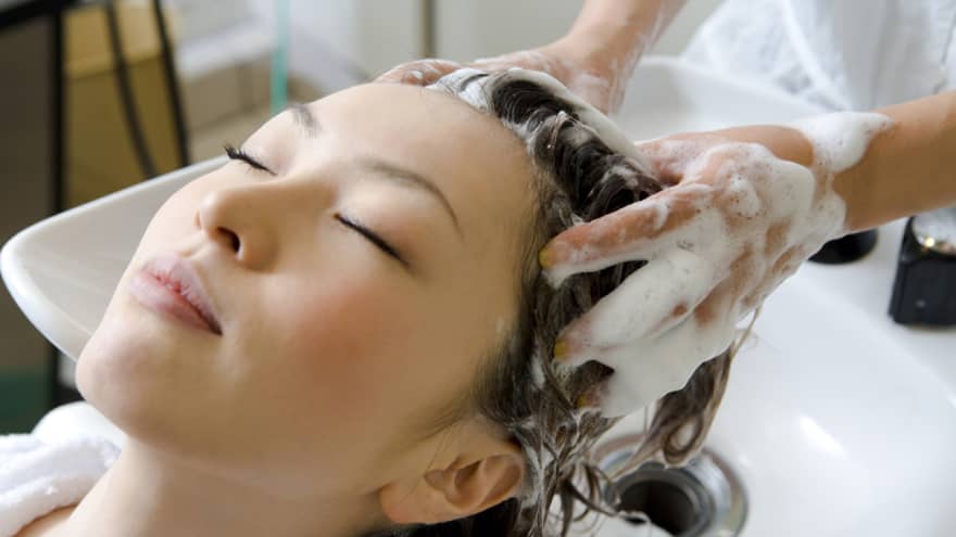 Woman closes eyes and leans back against sink as hands massage shampoo into her scalp