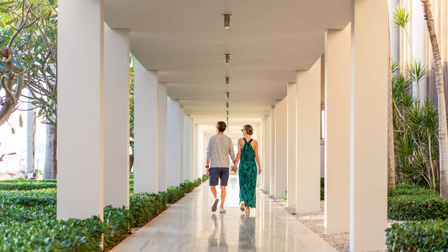 A couple in dressy attire walks hand-in-hand down an outdoor walkway lined with white columns