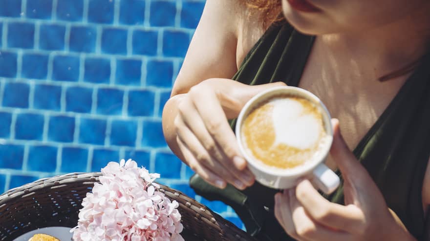 Overhead shot of swimsuit-clad woman sipping cappuccino in the pool, a tray of food in front of her