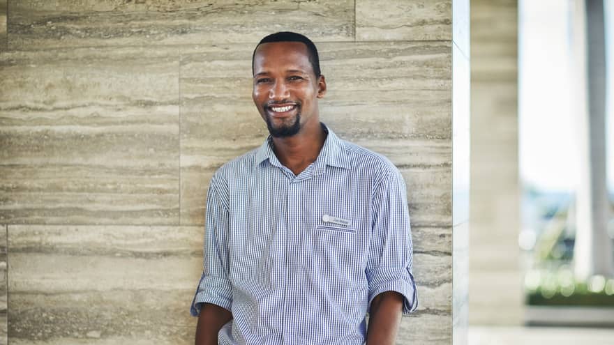 Smiling person in a checkered shirt standing against a stone wall indoors.