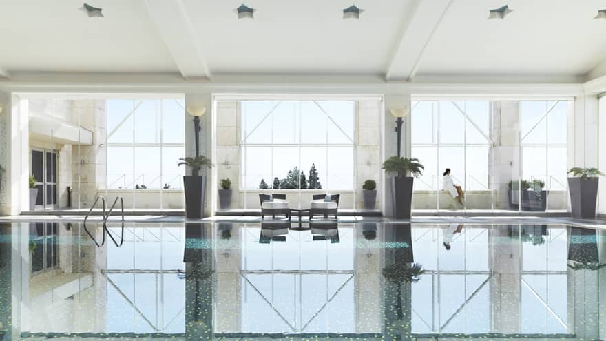 Indoor swimming pool reflecting three tall windows, woman in white bathrobe sits on ledge