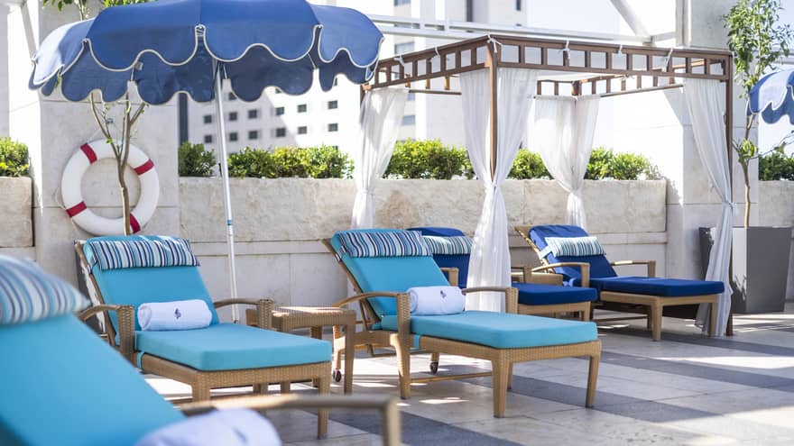 Wooden lounge chairs with team cushions, a pergola with white curtains drawn back and a blue scallop-edge umbrella on a pool deck