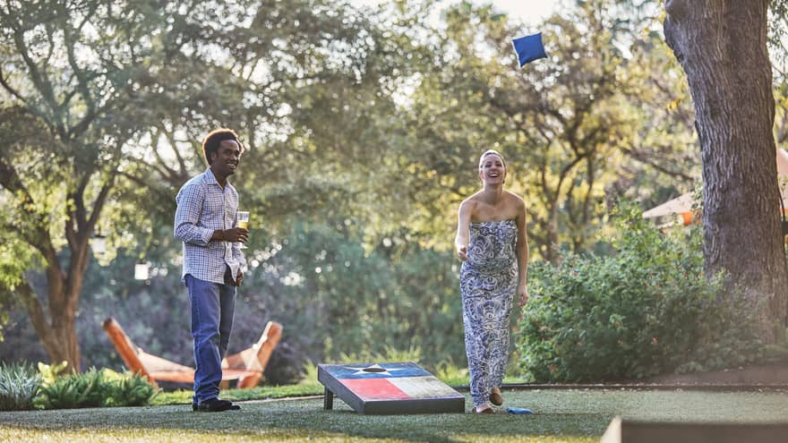Laughing woman tosses bean bag on lawn, man holding beer watches
