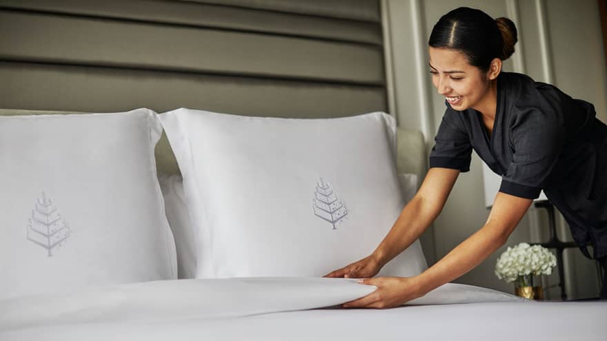 Hotel housekeeper smooths blanket under bed pillows with embroidered Four Seasons logos