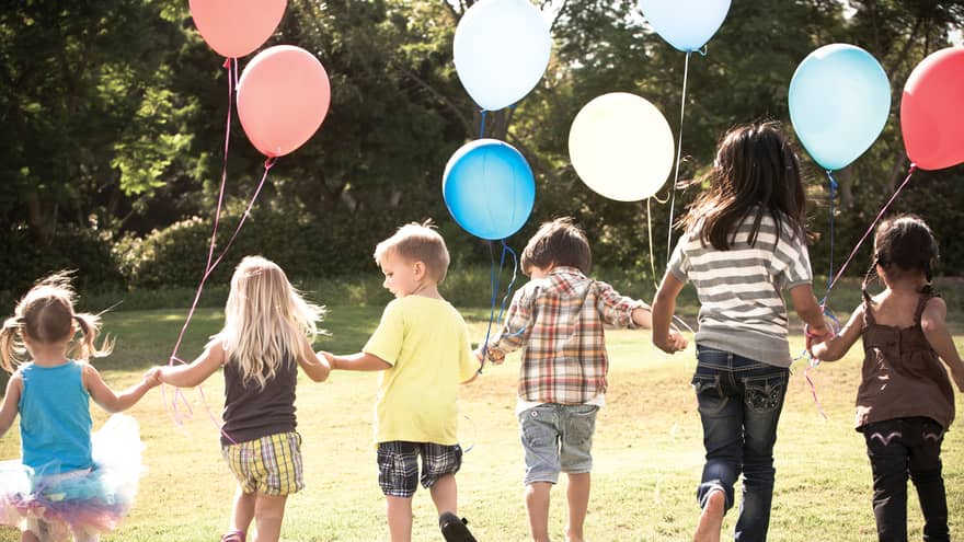 Children walking outside in the sunshine, holding hands, carrying coloured balloons.