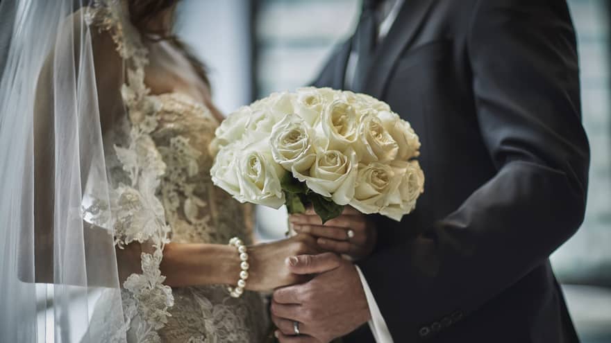 A close up of a bride in a lace gown and a groom in a gray suit hold the bride's bouquet of white roses together.