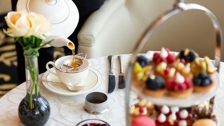 Table with afternoon tea service, tray with small sweets and pastries, rose in vase 