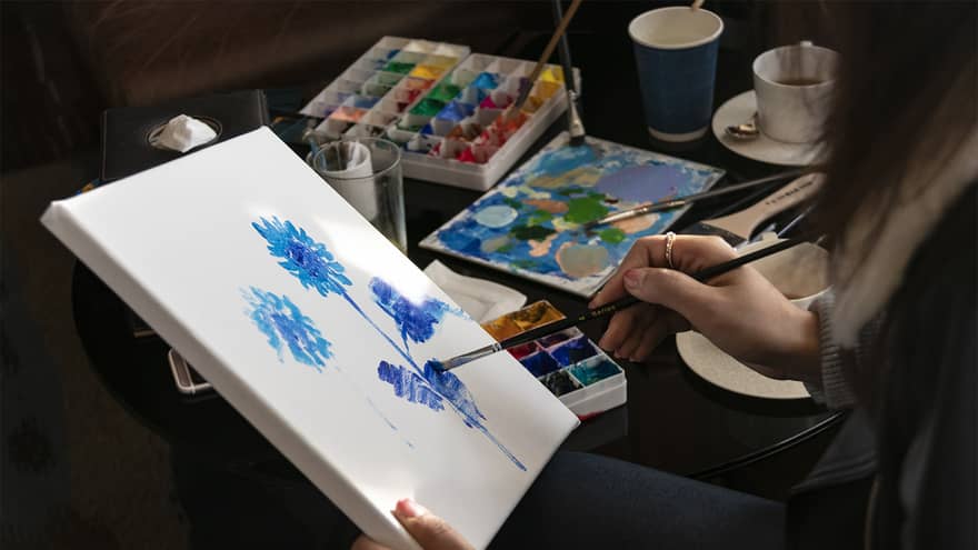 Close-up of woman painting blue flowers on white canvas 