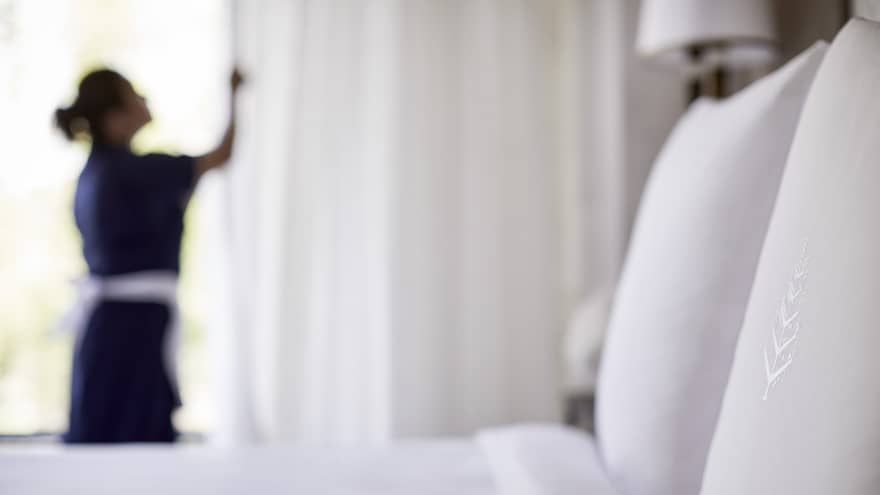 Close-up of bed, pillow with silhouette of hotel staff in uniform closing white curtains in background