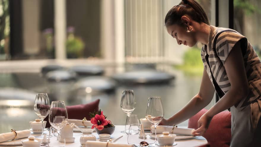 Woman sets a round table at restaurant, looking out to a tranquil pond