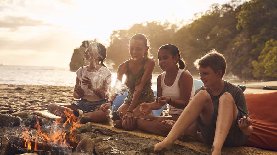 A group of young kids on a beach sitting near a small fire.