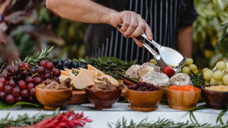 A chef's hand holds silver tongs over a display of grapes, dried fruits and cheese slices