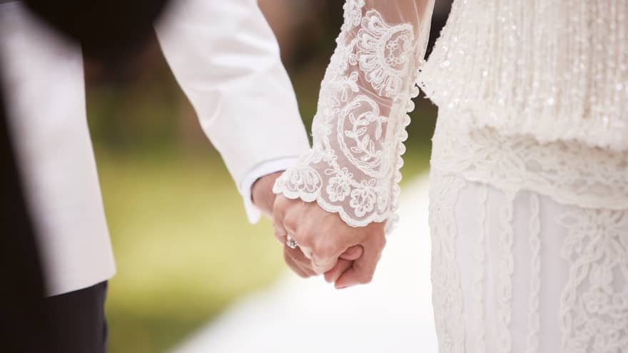 Close-up of bride with lace sleeve and groom with white suit holding hands 