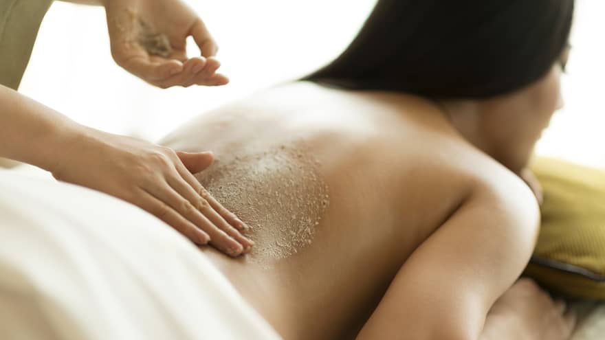 Woman laying face-down on massage table under white sheet as woman massages her back with cream
