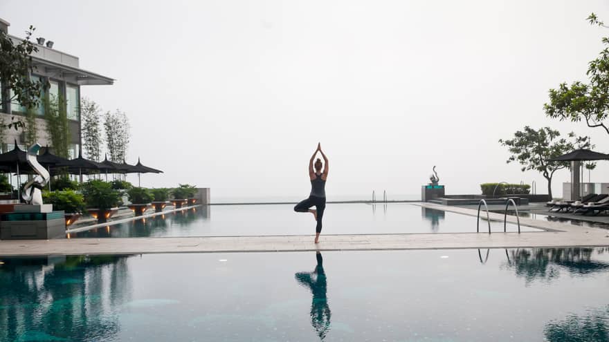 A person practices yoga, standing on one leg in a tree pose by a tranquil pool with modern landscaping and outdoor seating under umbrellas.