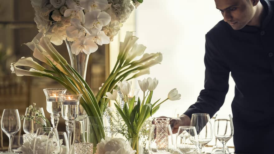 Hotel staff sets elegant banquet dining table with crystal glasses, vases with white flowers
