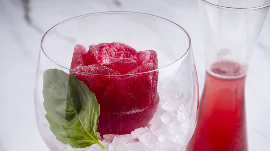 A frozen red rose?shaped creation garnished with a leaf, served on ice in a clear glass, with a nearby carafe of red beverage.