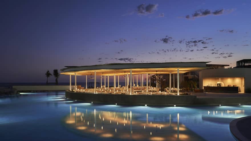 The exterior of an outdoor eating area at night on the water of a shore, it is well lit with lights.