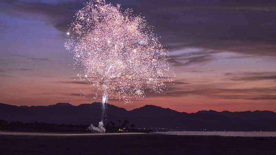 Pink fireworks exploding during sunset with a mountain behind them.