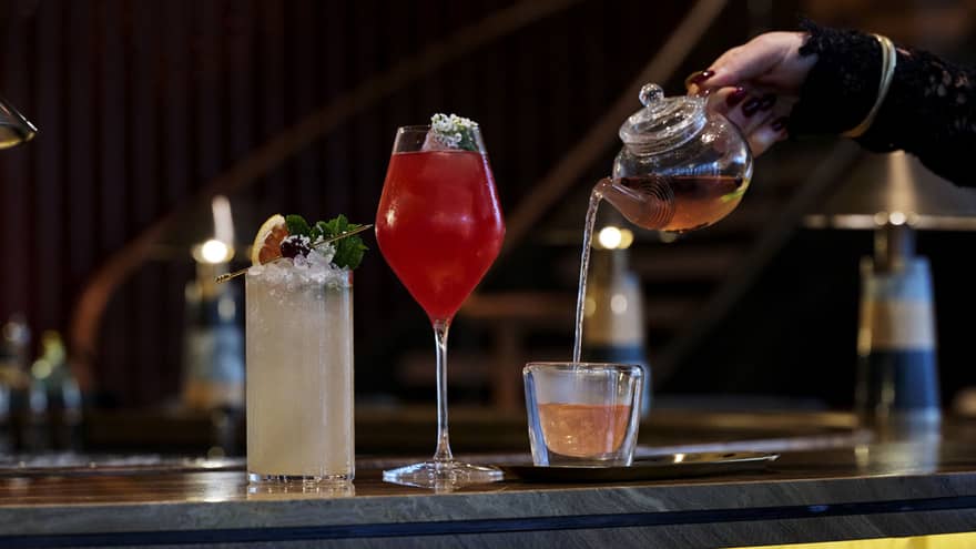 Two cocktails shown on a bar next to another beverage being poured into a glass from a small teapot