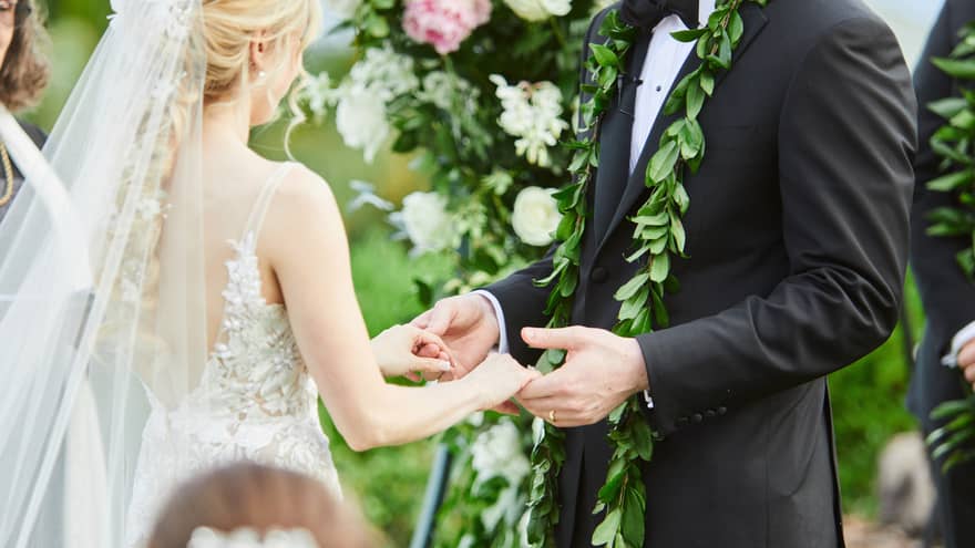 A couple saying their vows beneath a large floral arch. The woman has a chapel length veil and a lacy dress, the man is wearing a black suit with a floral garland around his neck.