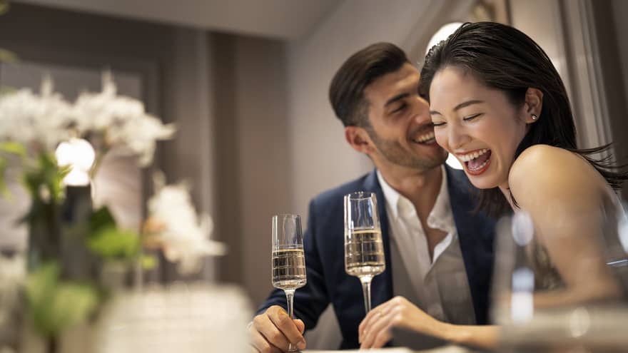 A couple sharing a meal on a table adorned with white flowers, holding champagne glasses and engaged in conversation and laughter.