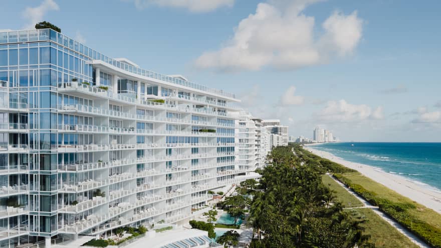 Luxury beachfront hotel with modern glass architecture, lush greenery, a pool area and a view of the ocean and sandy beach under a blue sky