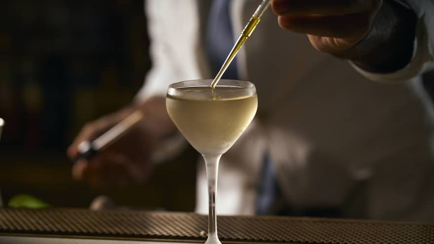 A hand uses a pipette to top up a gold-coloured martini in a stemmed glass with a rounded edge, in a darkly lit room.