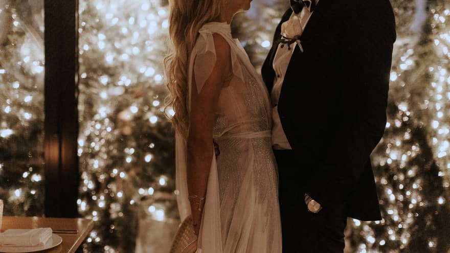 A stylish couple, dressed in elegant white and black, stand close, facing each other in a radiant wedding venue adorned with twinkling lights and greenery